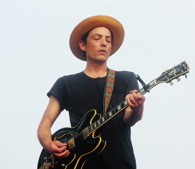 Jakob Dylan in a wide-brimmed hat playing a black hollow-body guitar on stage