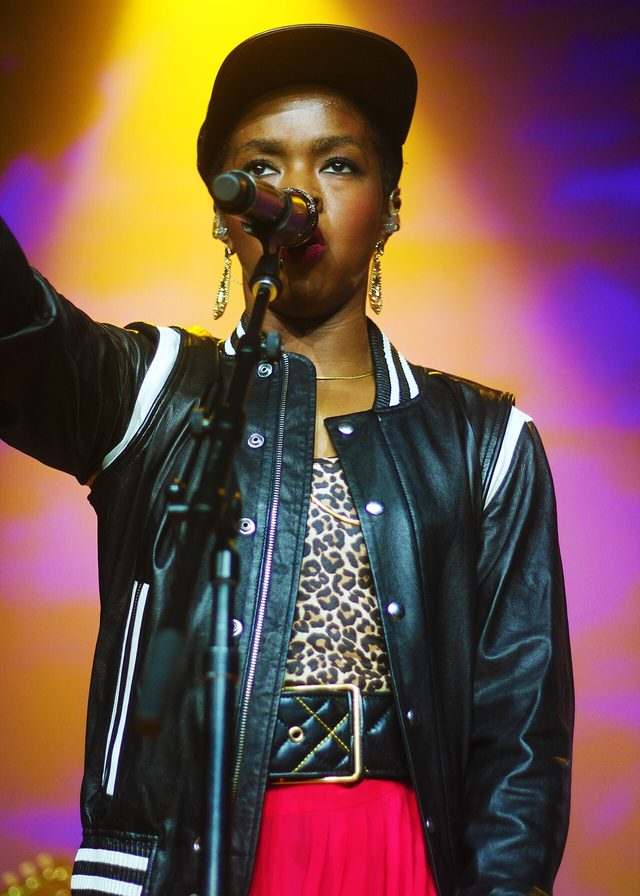 Ms. Lauryn Hill onstage in a black varsity jacket and cap, singing into a microphone