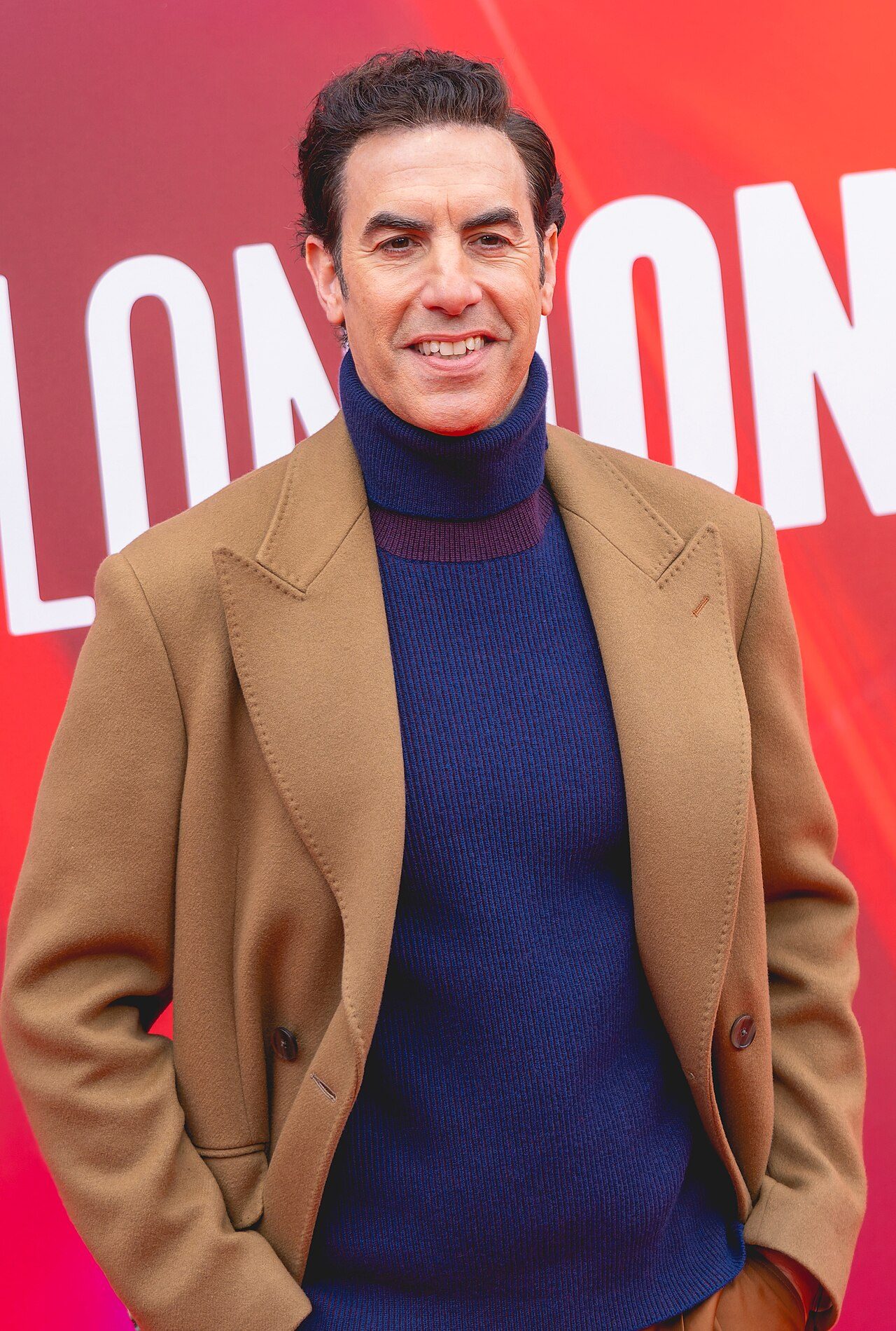 Sacha Baron Cohen smiling in a camel coat and navy turtleneck on a red carpet in London in 2024