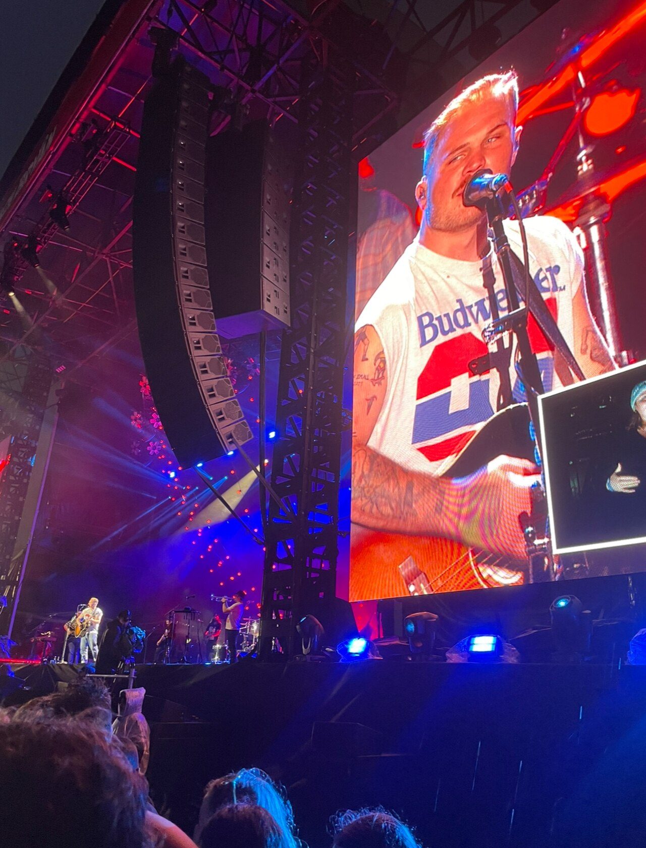 Zach Bryan in a white T-shirt singing at a microphone, seen on a big screen above a concert stage in Melbourne