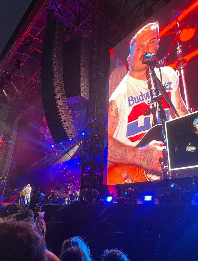 Zach Bryan in a white T-shirt singing at a microphone, seen on a big screen above a concert stage in Melbourne