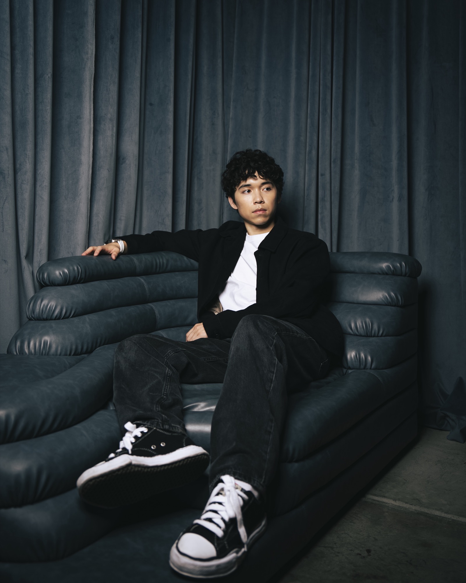 Asher Lara seated on a dark leather sofa against gray curtains, looking off to the side