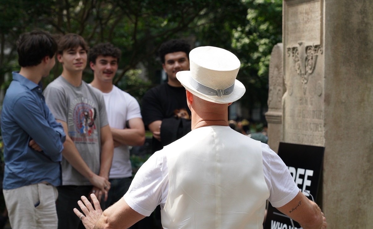 David Drebin, in a white hat and vest, talks with a group of young men in Washington Square Park