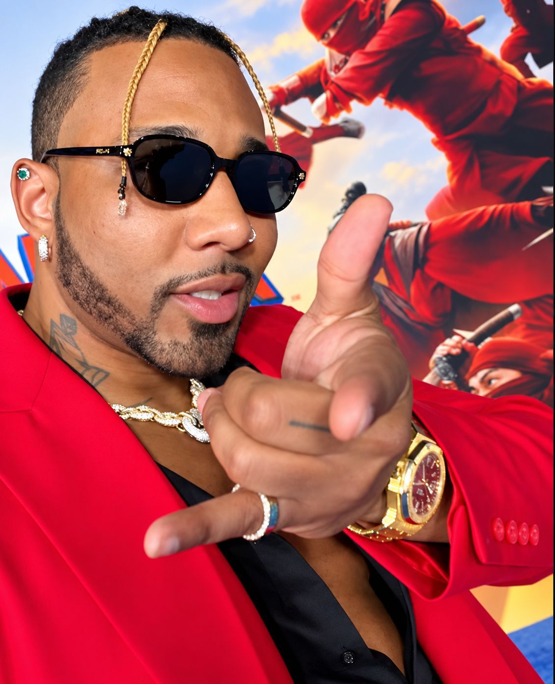 Eren Legend in a red blazer, sunglasses and gold watch, pointing at the camera in front of a Spider-Man premiere backdrop